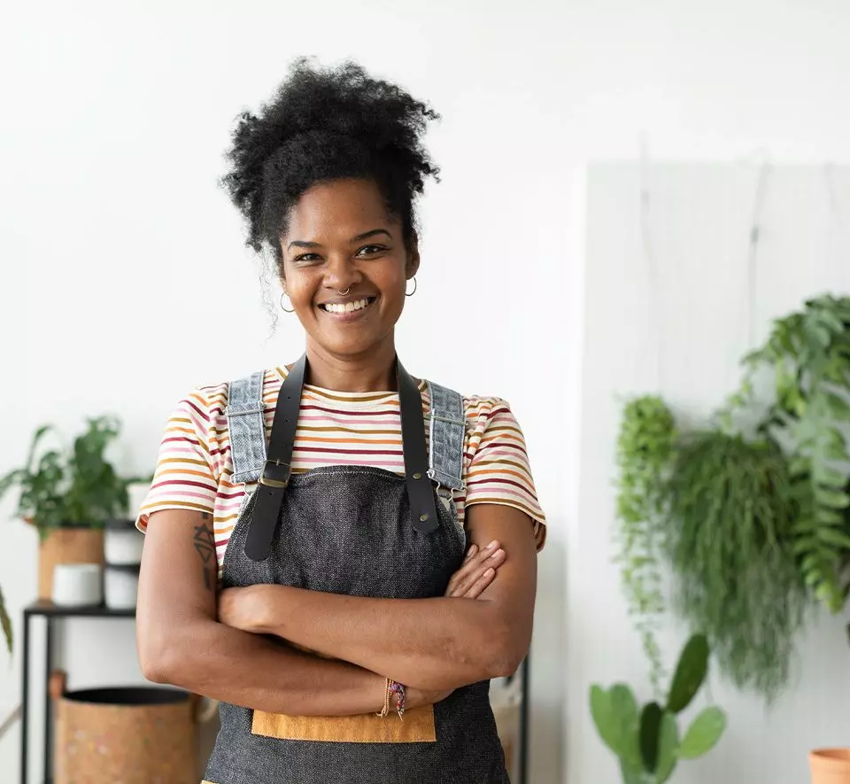 Avo Vision Woman with curly hair wearing a striped shirt and apron stands with arms crossed, smiling in a room with plants in the background, ready to lead an engaging training session.