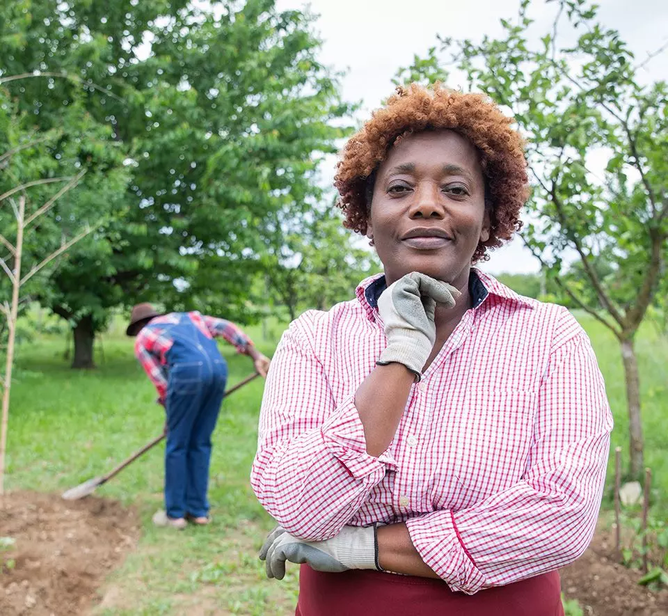 Avo Vision A person in a checkered shirt and gardening gloves stands with a hand on their chin, contemplating like an innovator, while another person in overalls rakes the ground in the background. Trees and greenery surround them.